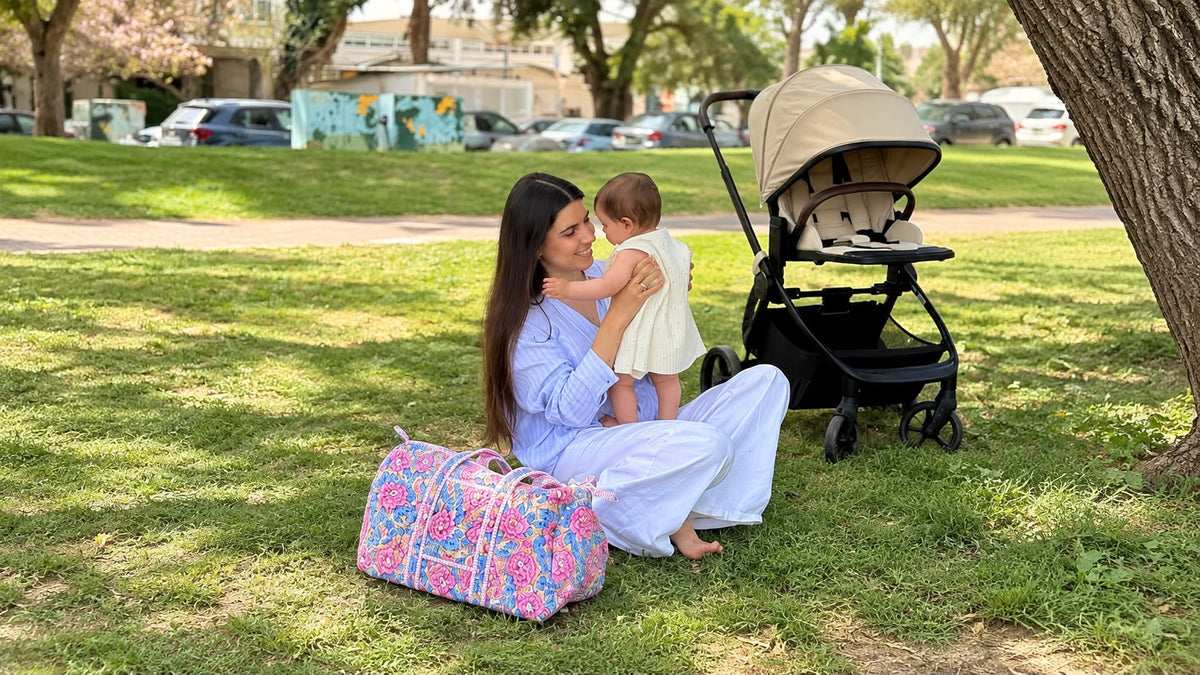 אם ותינוקת על הדשא בפארק, לצידן תיק ספרא פרחוני ועגלה